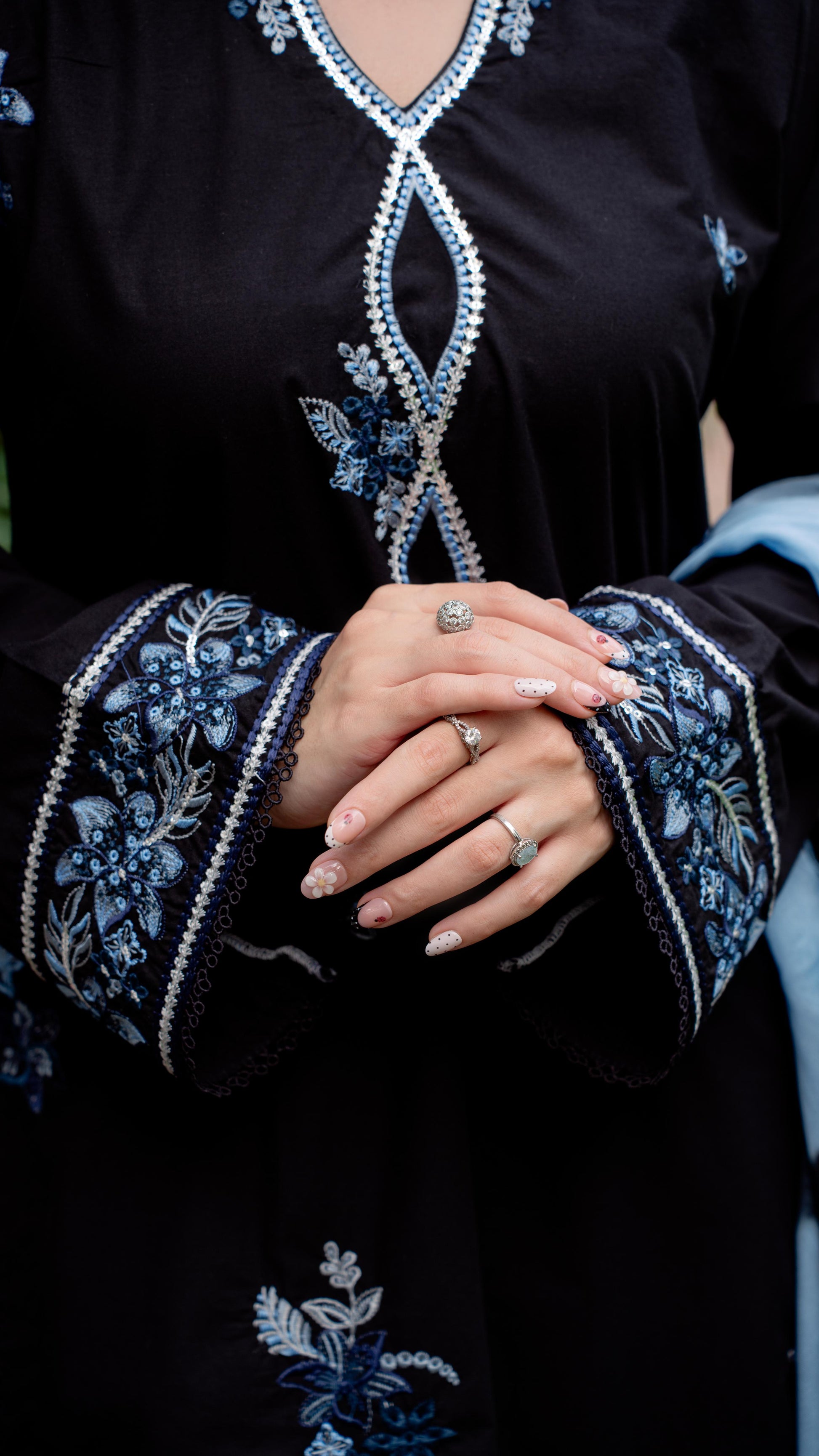 A close-up of a black embroidered dress or kurta with blue floral embroidery and silver beaded trim along the neckline, cuffs, and hem, worn by a model whose hands are clasped in the center.